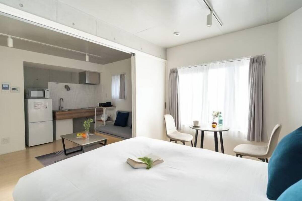 Une chambre d’hôtel avec un lit, une petite table avec une plante, une chaise, un coin cuisine et une fenêtre avec des rideaux.