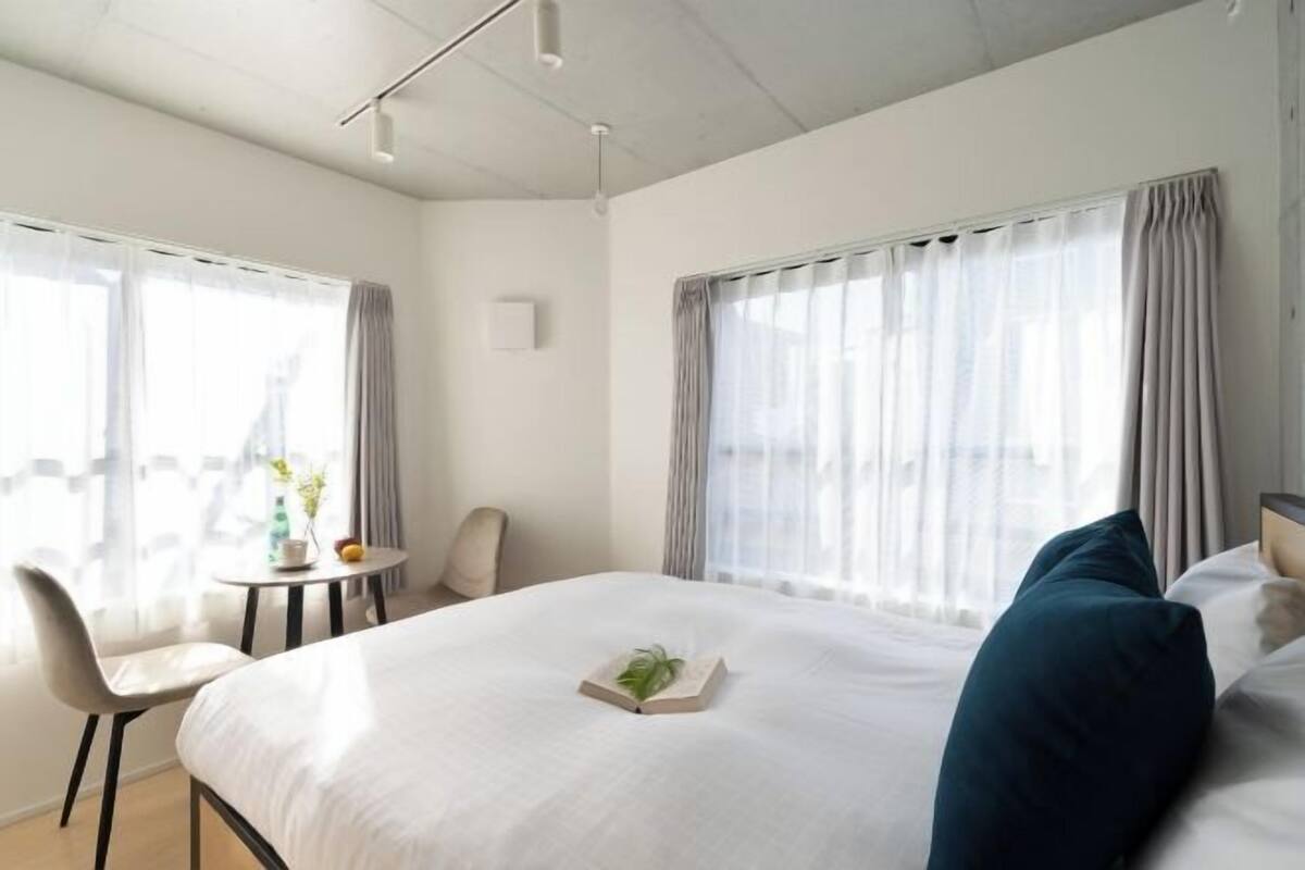 A hotel room with a bed, a chair, a small table with a plant, and large windows with curtains.