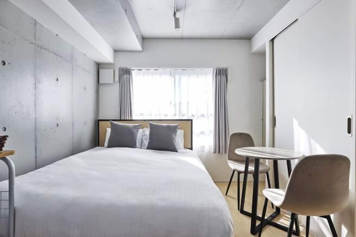 A modern hotel room with a large bed, a small table, two chairs, and a window with curtains.