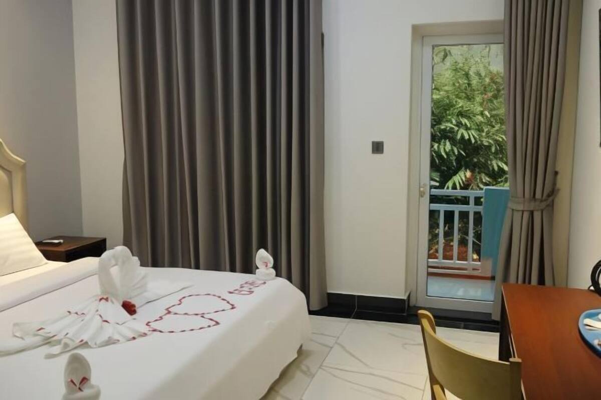 A hotel room with a bed, a small table, a chair, a balcony with a view of greenery, and a window with curtains.