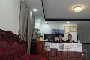 A reception desk with two staff members, a red patterned sofa, and a ceiling light fixture. - Suorsdei Sawadee (Kampot)