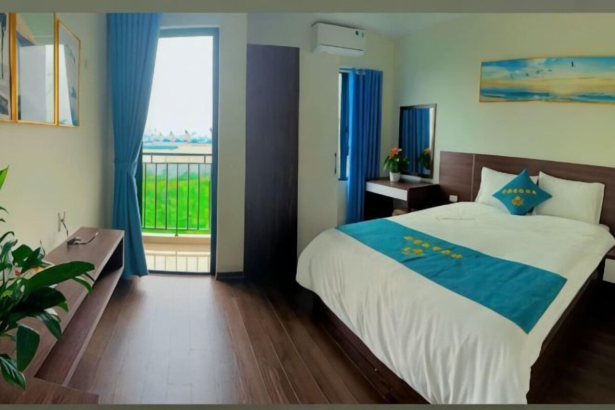 A hotel room with a bed, a desk, a chair, a potted plant, and a balcony with a view.