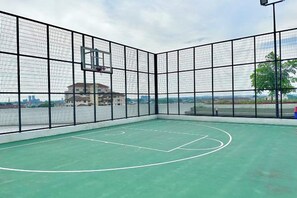 An indoor basketball court with a net, surrounded by a black mesh fence, and a view of buildings in the background.
