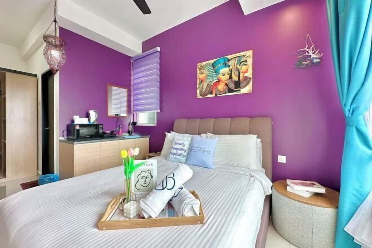 Quarto de hotel com cama, mesa de cabeceira, persianas e parede roxa.