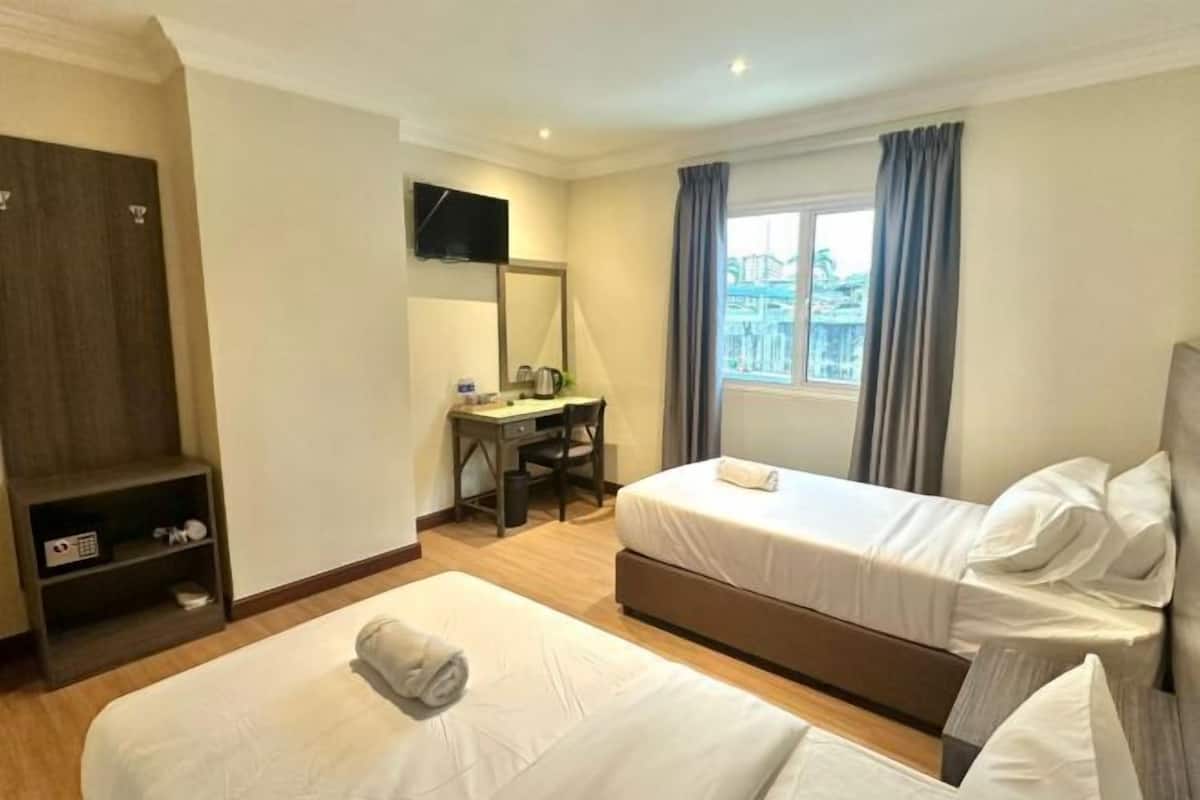 A hotel room with two beds, a desk, a TV, a window with curtains, and a small shelf with items.