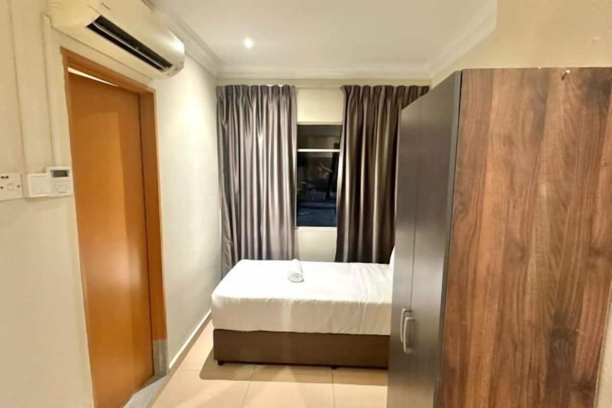 A bedroom with a bed, a wooden wardrobe, a door, and an air conditioning unit.