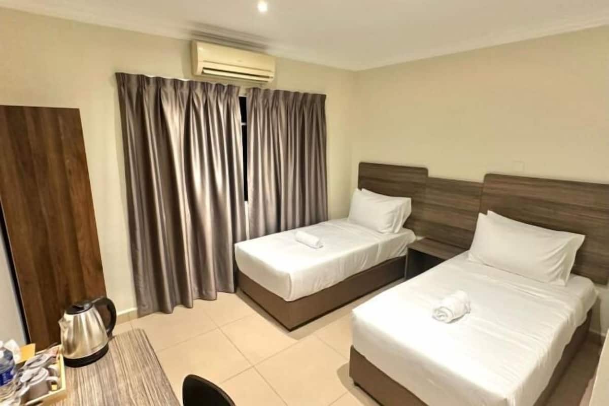 A hotel room with two beds, a wooden headboard, a small table with a kettle, and a curtain.