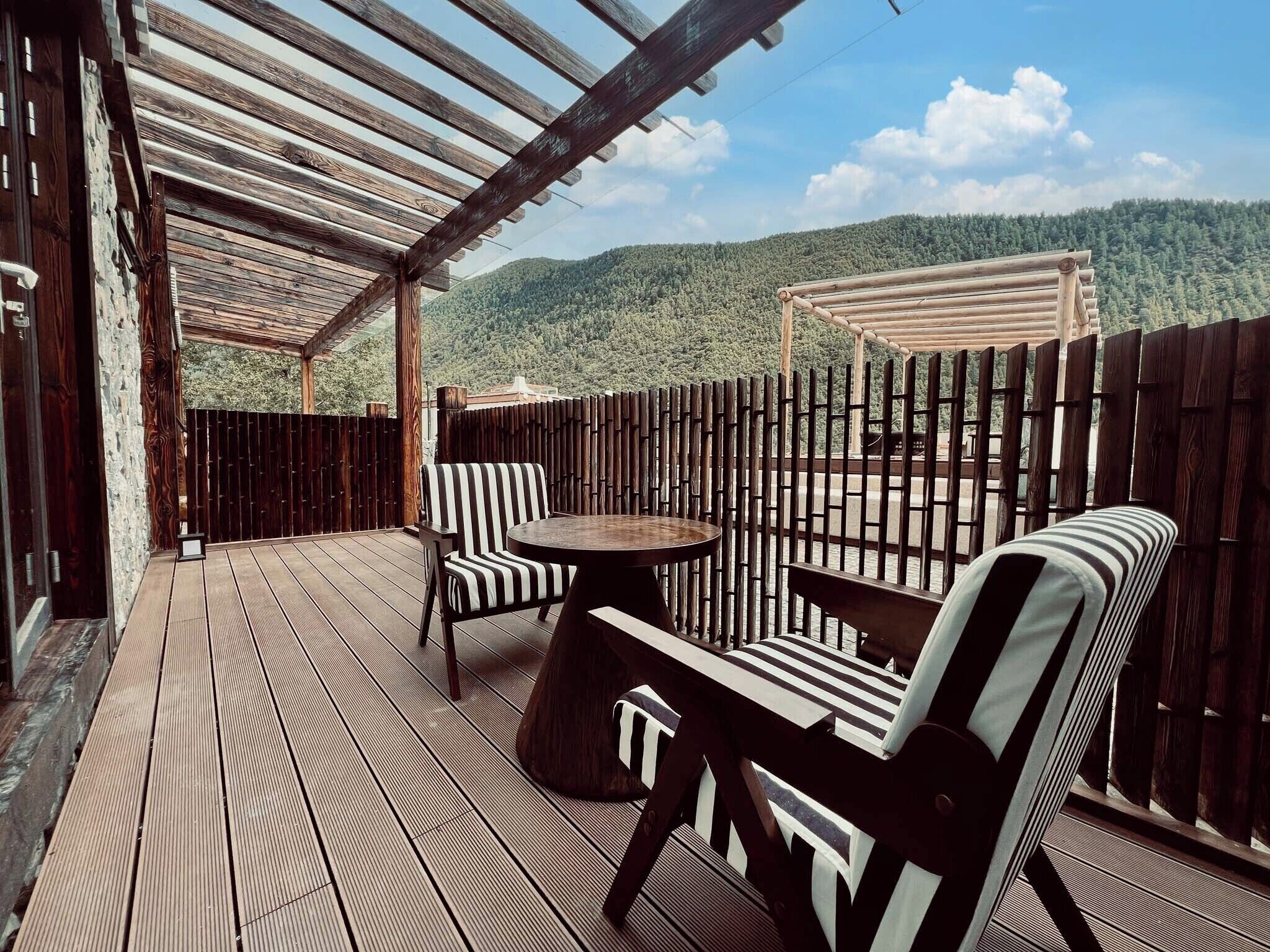 A wooden deck with a pergola, two striped chairs, and a round table.