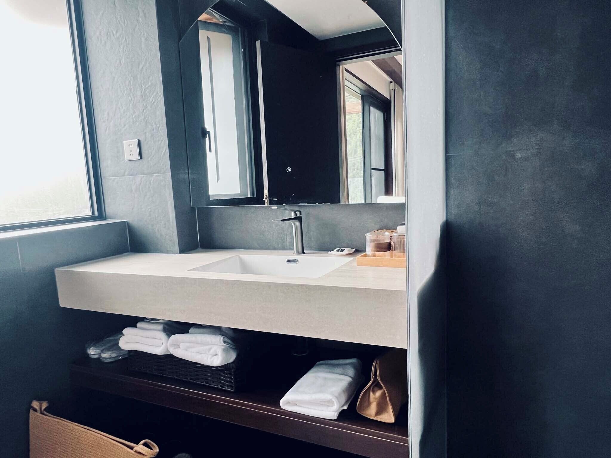 A modern bathroom with a large mirror, a sink, and a shelf with towels and toiletries.
