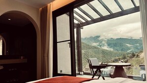 A hotel room with a large bed, a dining table with chairs, and a view of mountains through a large window. - Mid Hill White Lodge (Ngawa Autonomous Prefecture)