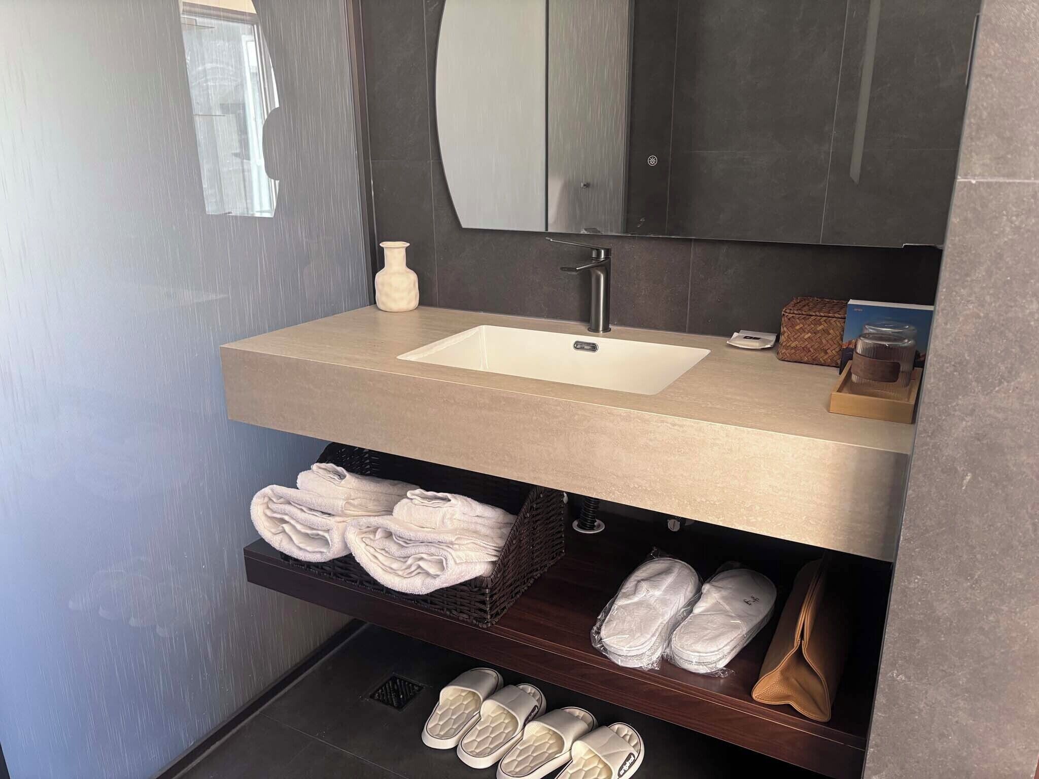 A bathroom with a beige sink, a mirror, a towel rack with folded towels, and a shelf with shoes and slippers.