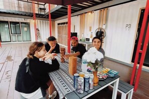 Un groupe de personnes est assis à une table en plein air, devant une variété de plats et de boissons. On aperçoit un ventilateur et, sur le mur derrière eux, un panneau portant le numéro « P3 ».