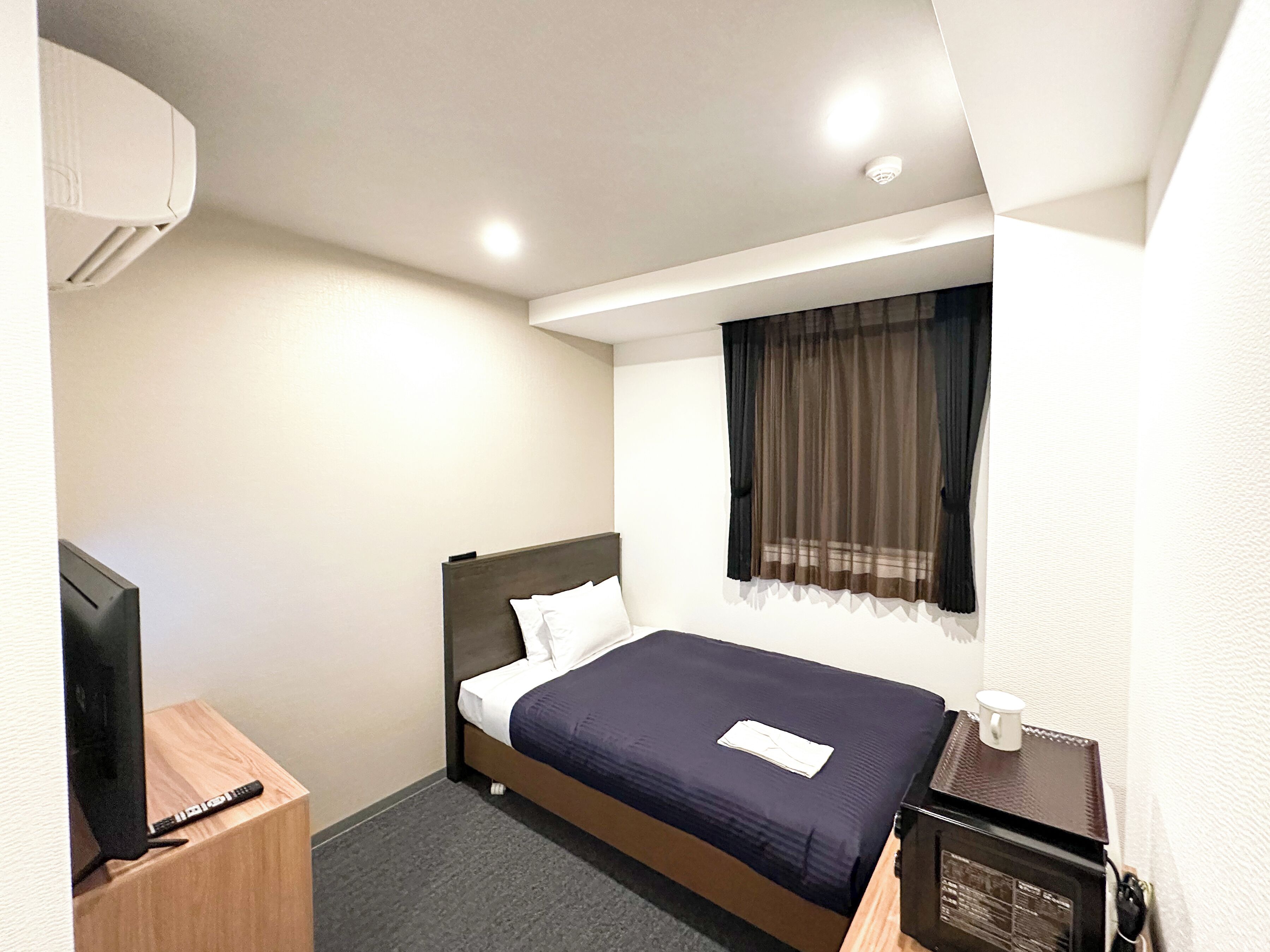 A hotel room with a single bed, a bedside table with a remote control, a small table with a cup, and a wall-mounted air conditioner.
