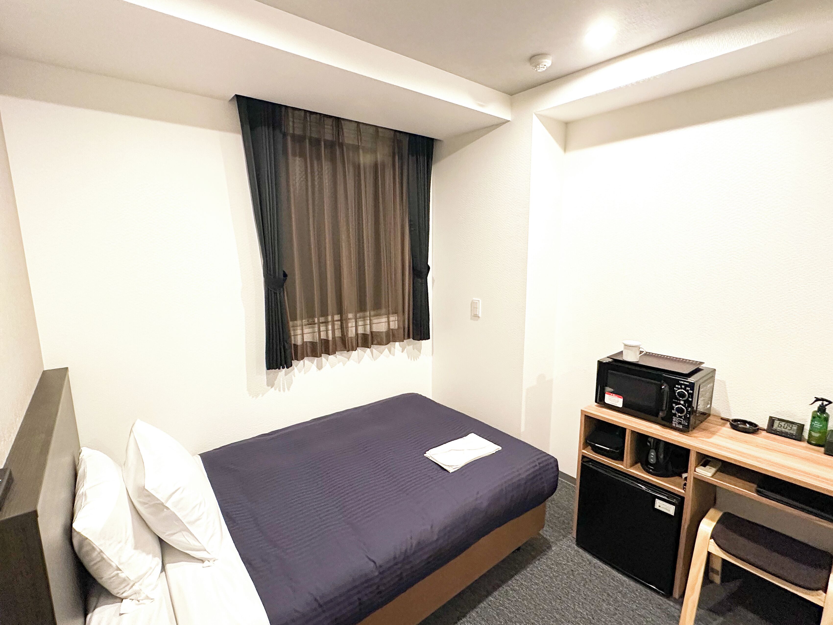A small, neatly arranged hotel room with a single bed, a desk with a microwave, a small refrigerator, and a window with sheer curtains.