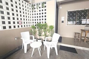 Un espace détente avec des chaises blanches et une petite table, une plante en pot et une fenêtre ornée d’une grille métallique.