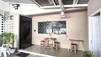 A covered outdoor area with a table and chairs, a small stool, a window with a grid pattern, and a door.
