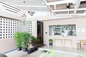 An indoor space with a ceiling with exposed beams, a white wall with a grid pattern, a window with a view, and several potted plants. - Relax Inn DMK (Bangkok)