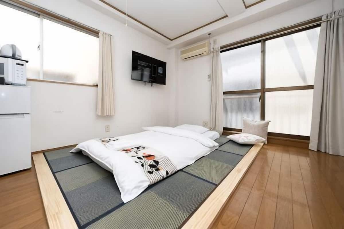 Um quarto com cama, pequeno espaço de cozinha e uma janela grande.