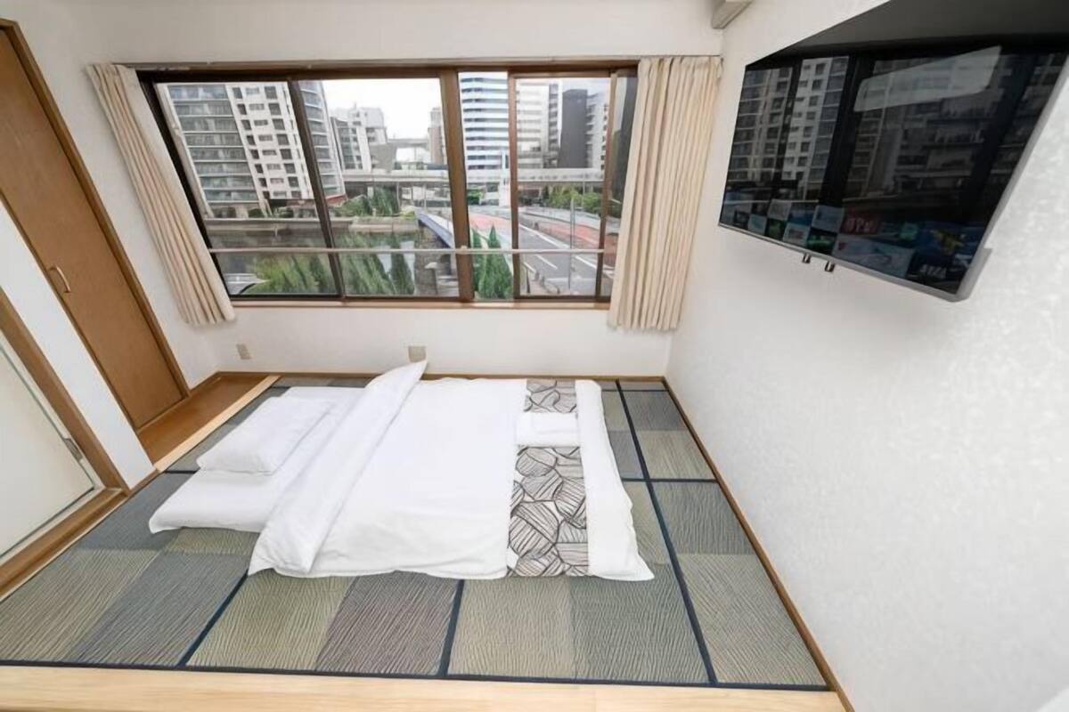 Quarto com piso de tatame, cama com roupa de cama branca, janela com vista para edifícios e tela fixada na parede.