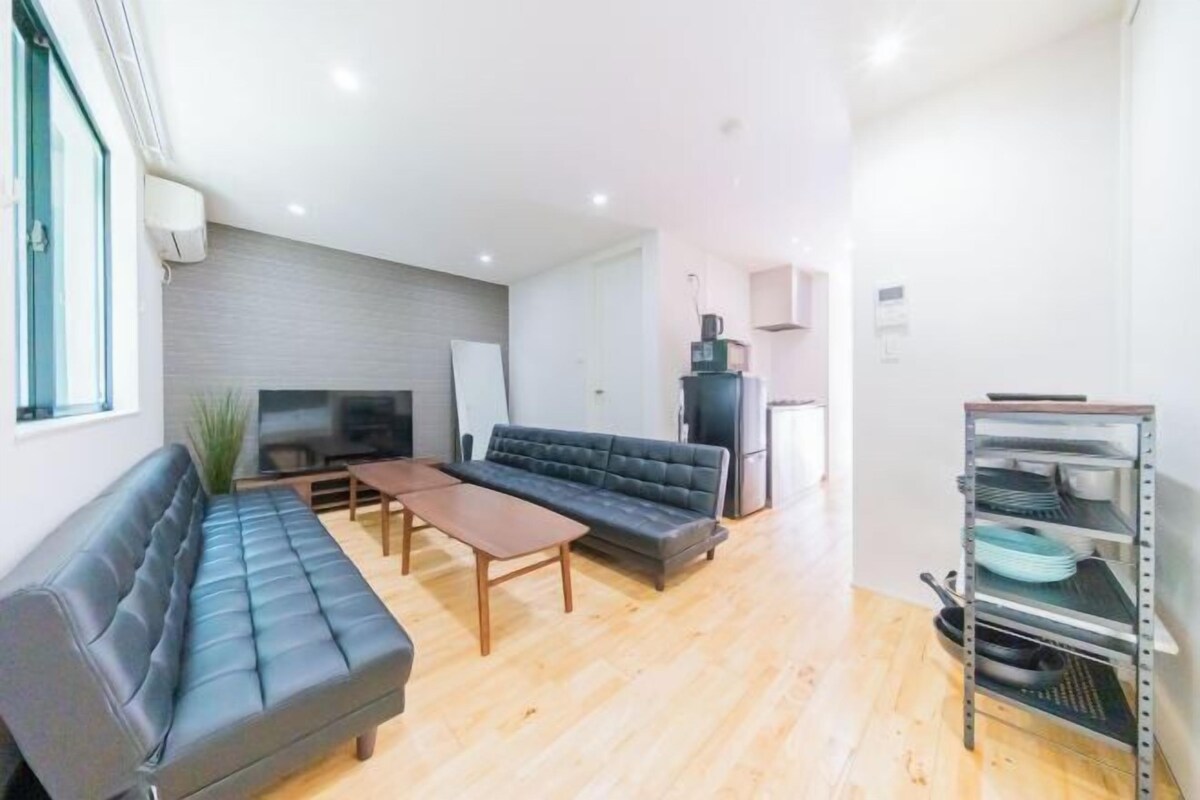 A modern living room with a sofa, a coffee table, a TV, and a kitchen area with a fridge and a shelf.