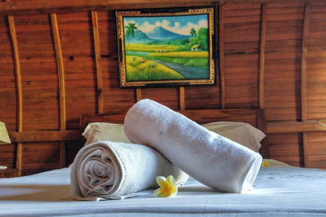 Una cama bien hecha con toallas enrolladas y una sola flor, un cuadro enmarco con un paisaje en la pared, y paneles de madera.