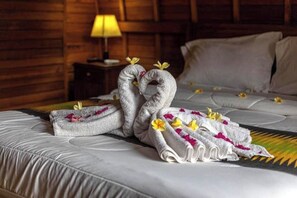 A neatly made bed with towels arranged in a heart shape, a lamp, and a wooden wall. - The Sweet Corn Bungalow (Bali)