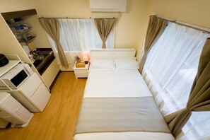 Hotellihuone, jossa on sänky, yöpöytä, lamppu, pieni pöytä, pesukone ja ikkuna, jossa on verhot.