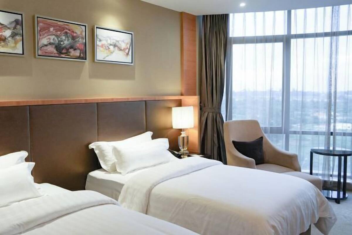 Une chambre d’hôtel avec deux lits, une chaise, une lampe et une vue sur la ville grâce à de grandes fenêtres.