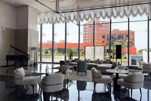 A hotel lobby with a grand piano, comfortable seating, and large windows offering a view of a red building.