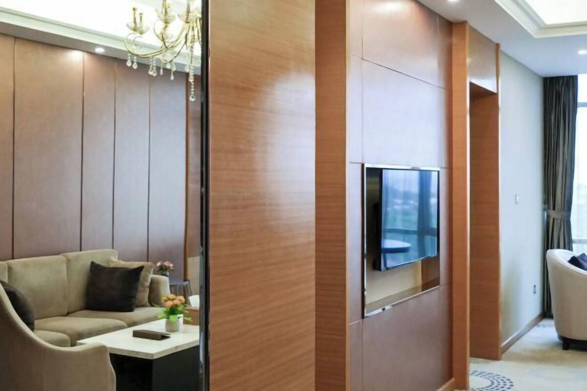 Une chambre d’hôtel moderne avec un canapé, un téléviseur fixé sur un panneau en bois et un lustre.
