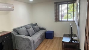 Habitació amb un sofà gris, un otomà blau petit, un armari de fusta, un escriptori amb un ordinador i una finestra amb persianes.