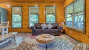 L’intérieur d’un chalet en bois, avec un canapé en cuir marron, deux fenêtres, un plafond en bois et un tapis à motifs.