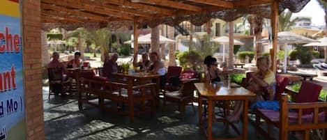 An outdoor patio with wooden tables and chairs, surrounded by greenery and umbrellas.