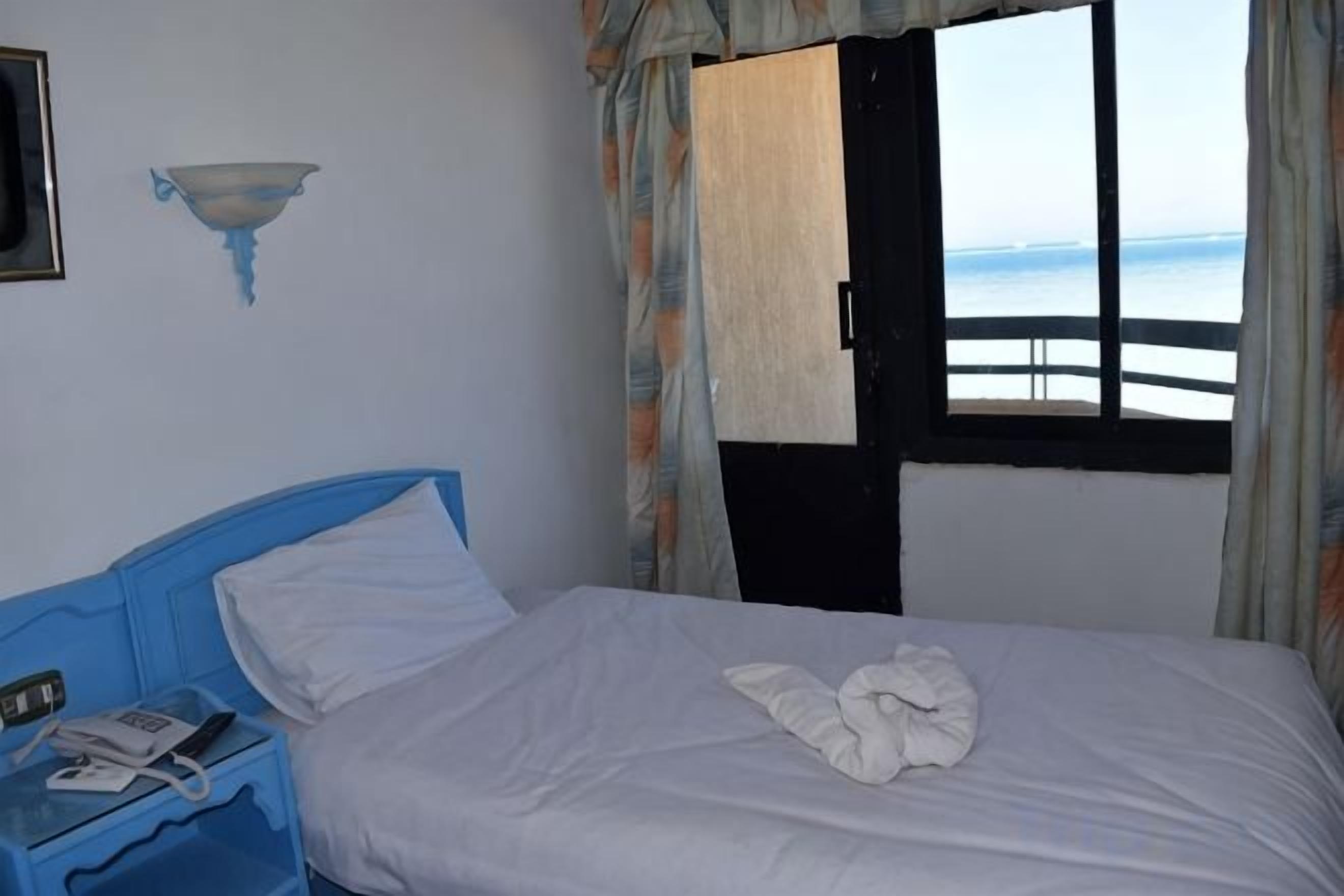 A bedroom with a bed, a blue headboard, a bedside table with a phone, and a window with a view of the sea.