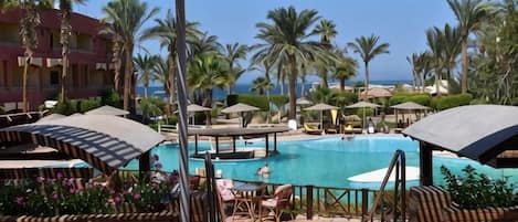 A pool area with lounge chairs and umbrellas, surrounded by palm trees and a view of the sea.
