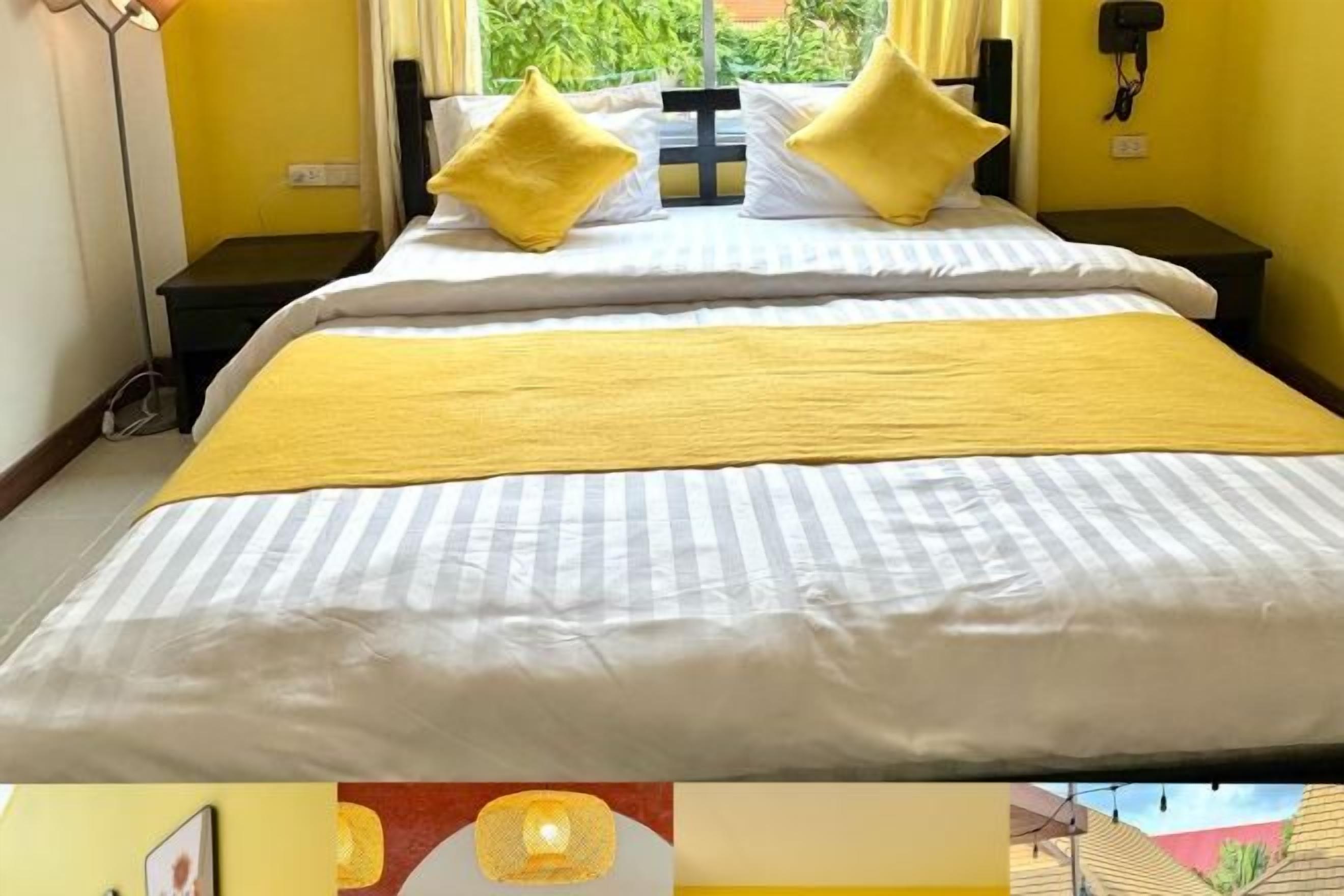 Habitación de hotel con cama, almohadones amarillos, mesita de noche y ventana con vistas a follaje.