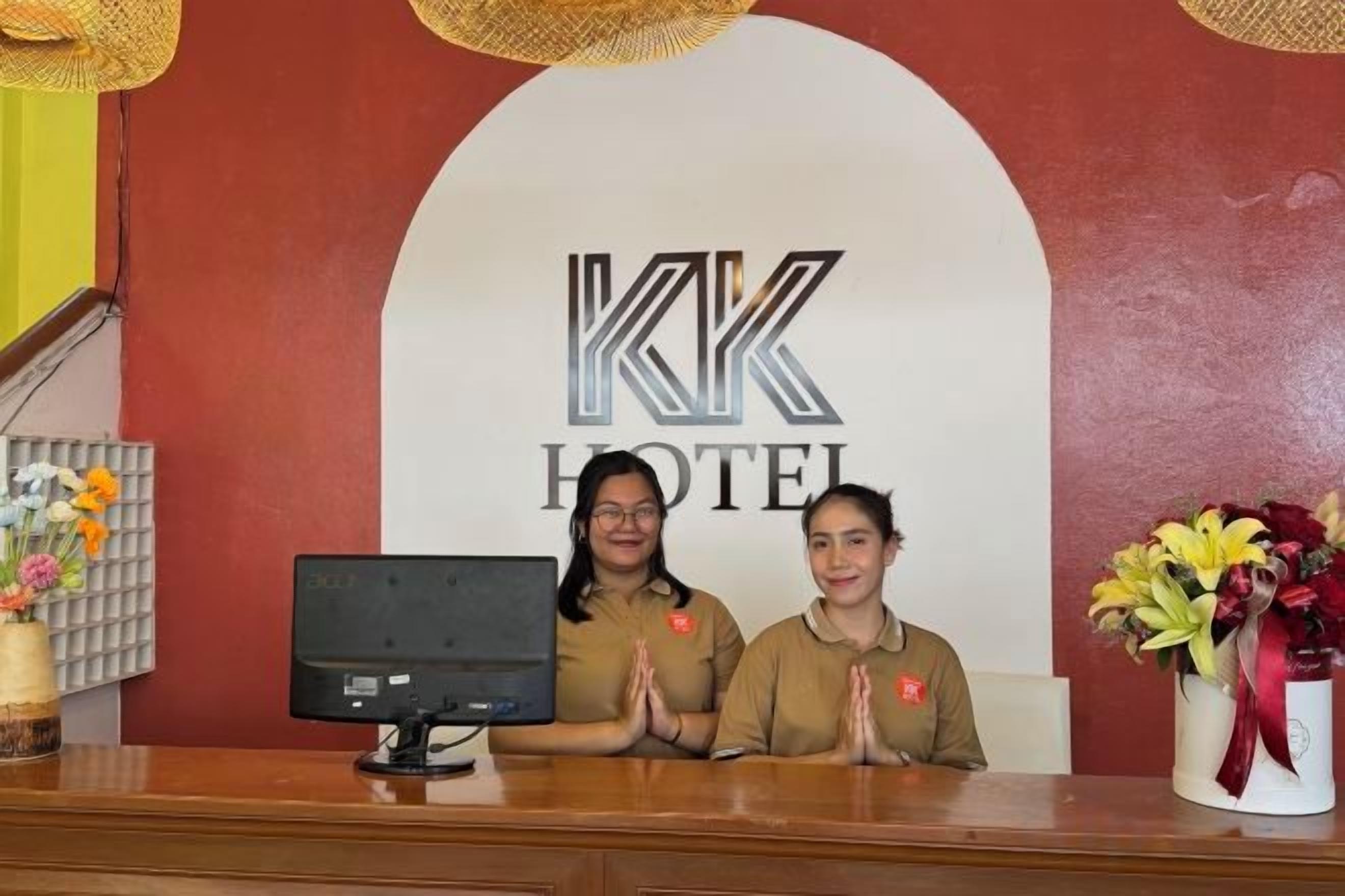 A hotel reception desk with a digital monitor, two receptionists, and a large hotel logo on the wall.