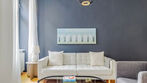 A modern living room with a white sofa, a grey armchair, a glass coffee table, and a painting on the wall.