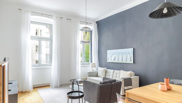 A modern living room with a grey sofa, a wooden dining table, and a large window with white curtains.