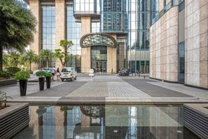 A modern building with a glass facade, a central entrance with a curved canopy, and a reflective water feature in the foreground. - Vinhomes Chic Apartment - Ho Chi Minh (Ho Chi Minh City)