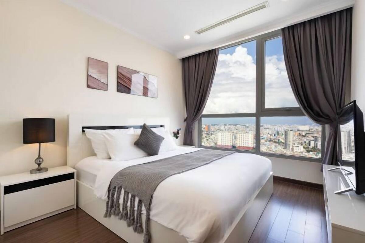 Ein Hotelzimmer mit Bett, Nachttisch, Lampe und einem großen Fenster mit Vorhängen.