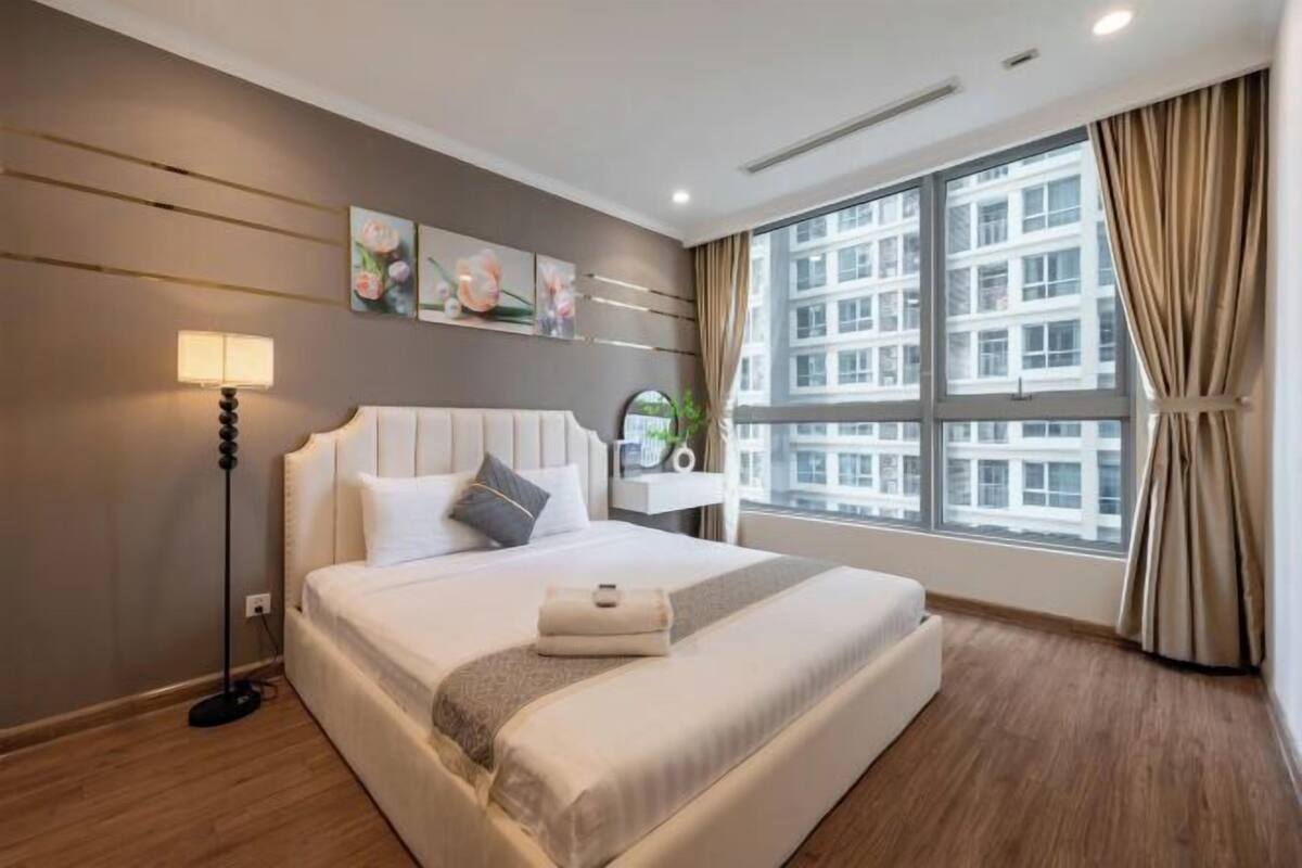 Ein Hotelzimmer mit einem großen Bett, einer Nachttischlampe, Blick auf Gebäude durch das Fenster und einem Wandbild.
