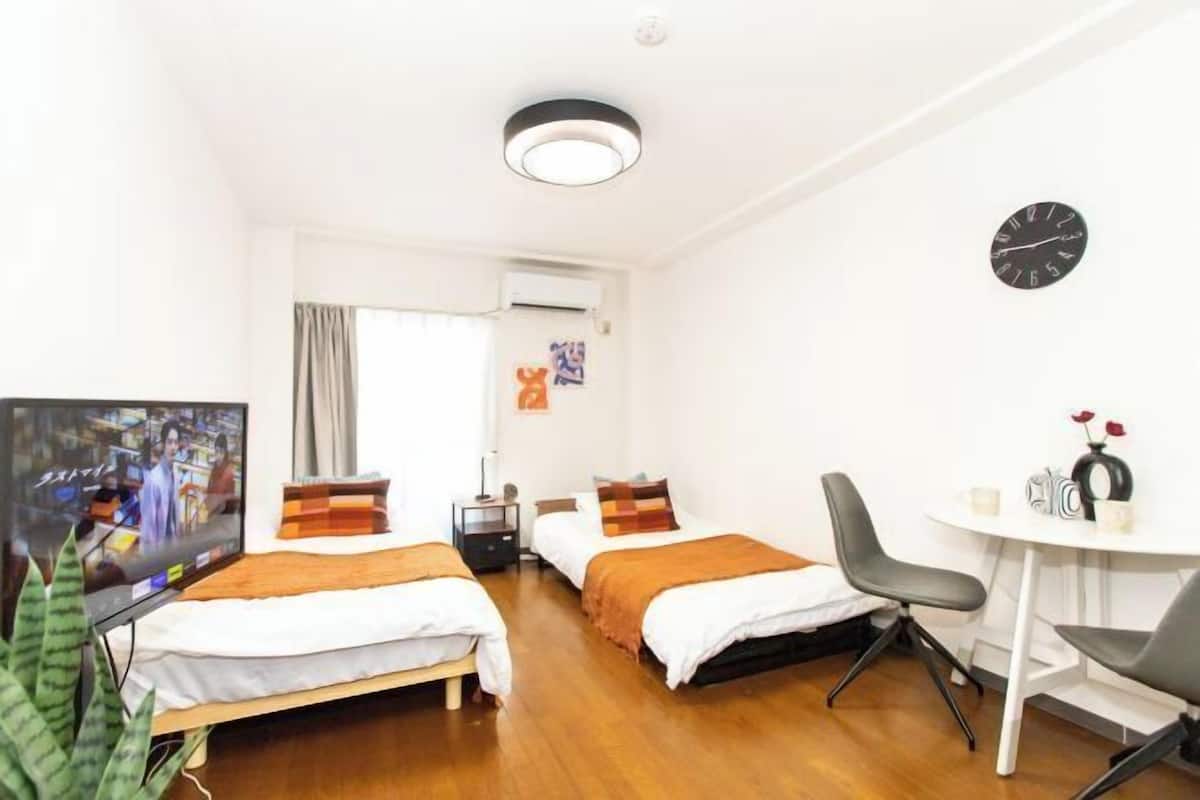 A hotel room with two beds, a TV, a desk with a chair, a clock, and a window with curtains.
