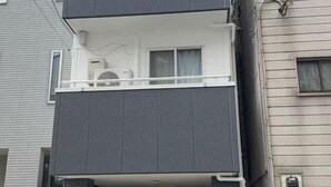 Un edificio de dos plantas con un balcón en el piso superior, que cuenta con una unidad de aire acondicionado pequeña y una ventana con cortina.