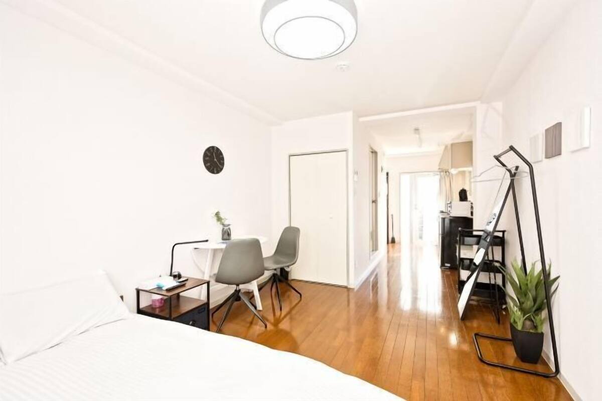 A hotel room with a bed, a desk, a chair, a small table, a clock, and a potted plant.