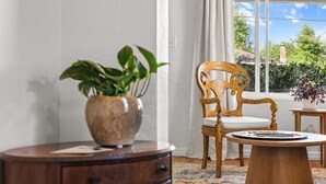 Una habitación con una silla de madera, una mesa redonda de madera, una mesita auxiliar de madera y una planta en maceta.
