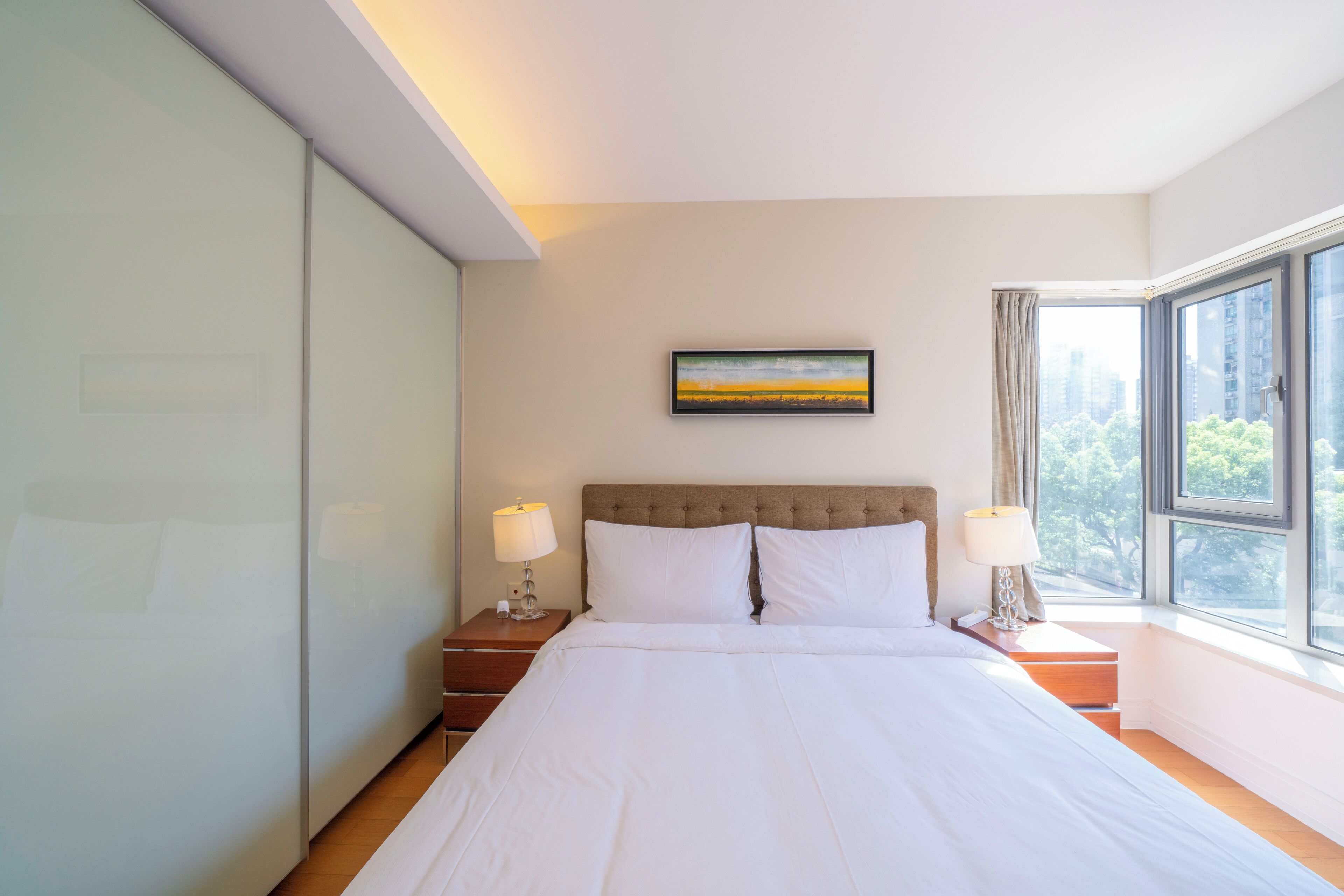 A hotel room with a large bed, a nightstand with a lamp, a painting on the wall, and a window with a view of buildings.