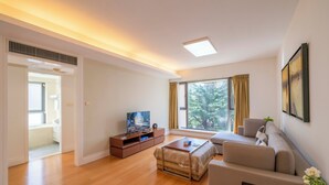 Un salon moderne avec un canapé, une table basse et un meuble pour la télévision. Il y a une grande fenêtre avec des rideaux, une porte donnant sur une autre pièce et un luminaire au plafond.