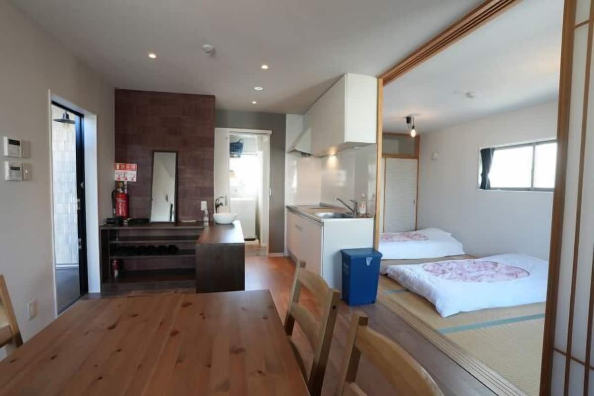 Ett kompakt hotellrum med ett matbord i trä, ett litet pentry, ett litet badrum och en säng med en futonmadrass.