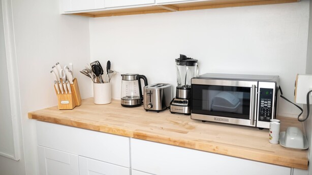 Una cocina con armarios blancos, encimera de madera, cafetera, tostadora, licuadora y microondas.